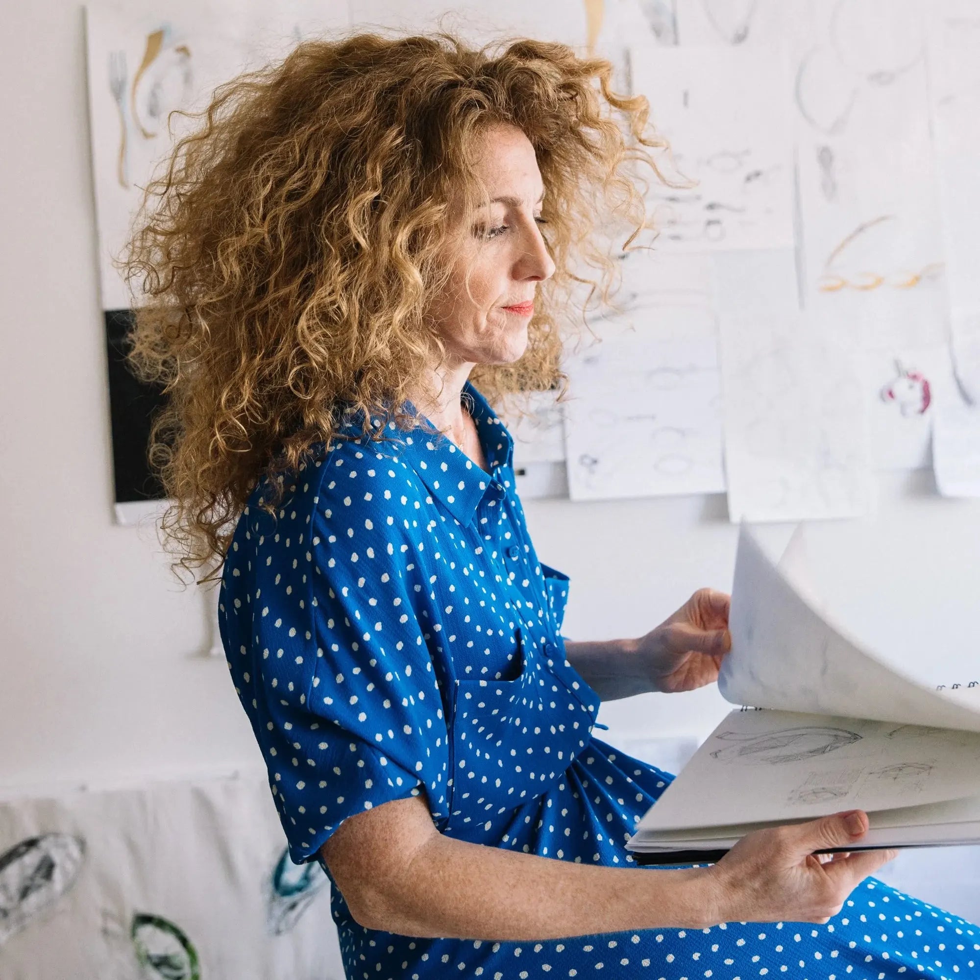 Woman, Ilaria Ilanzoni, in a blue polka dot dress sitting on a chair, holding a book in a room with white walls and artwork.