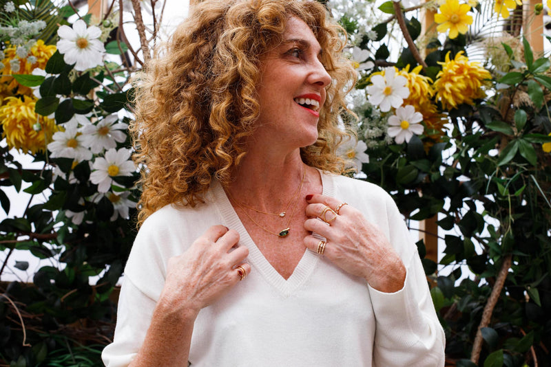 Woman with curly hair standing among flowers, wearing a white sweater and fabulous rings and necklaces.