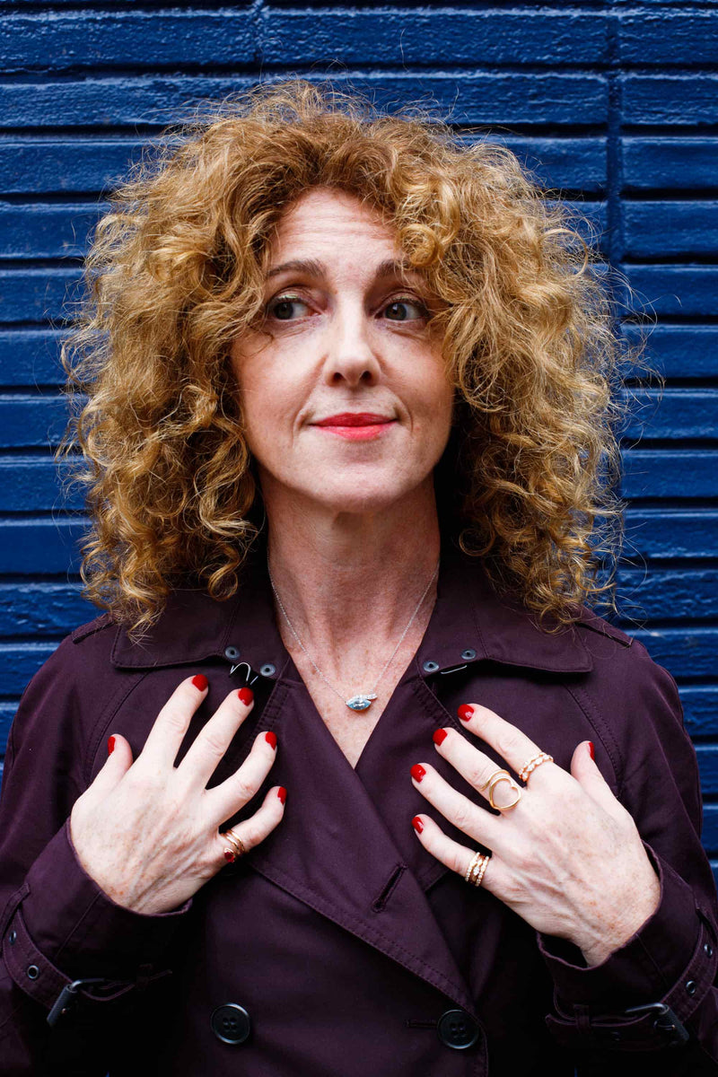 Woman with curly hair and red nails wearing a dark coat against a blue wall.