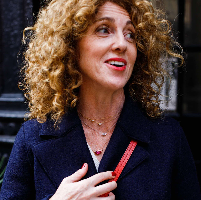 Woman with curly hair wearing a dark coat and red handbag, looking to the side.